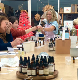 Group of women at an indoor DIY perfume workshop in a craft studio, sampling essential oil blends from amber dropper bottles while an instructor passes a bottle, pink holiday tree and paper star decorations in the background.