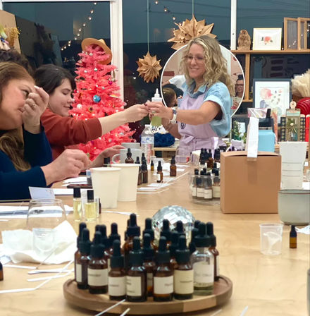 Group of women at an indoor DIY perfume workshop in a craft studio, sampling essential oil blends from amber dropper bottles while an instructor passes a bottle, pink holiday tree and paper star decorations in the background.