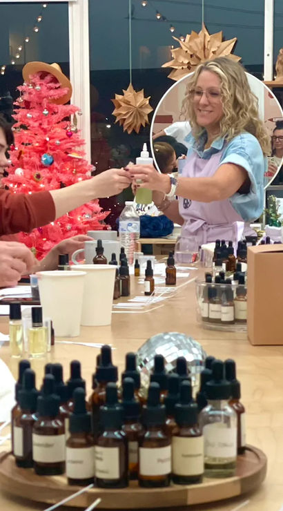 Group of women at an indoor DIY perfume workshop in a craft studio, sampling essential oil blends from amber dropper bottles while an instructor passes a bottle, pink holiday tree and paper star decorations in the background.