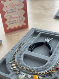 Close-up of a jewelry-making station: gold charm bracelet with colorful enamel and gemstone charms laid on a gray beading tray with numbered sizing marks, black pliers, and a blurred pricing sign in the background.