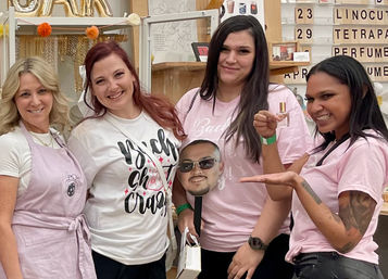 Four smiling women in a bright craft studio pose at a DIY perfume workshop — one holds a tiny fragrance bottle and another displays a playful face cutout.