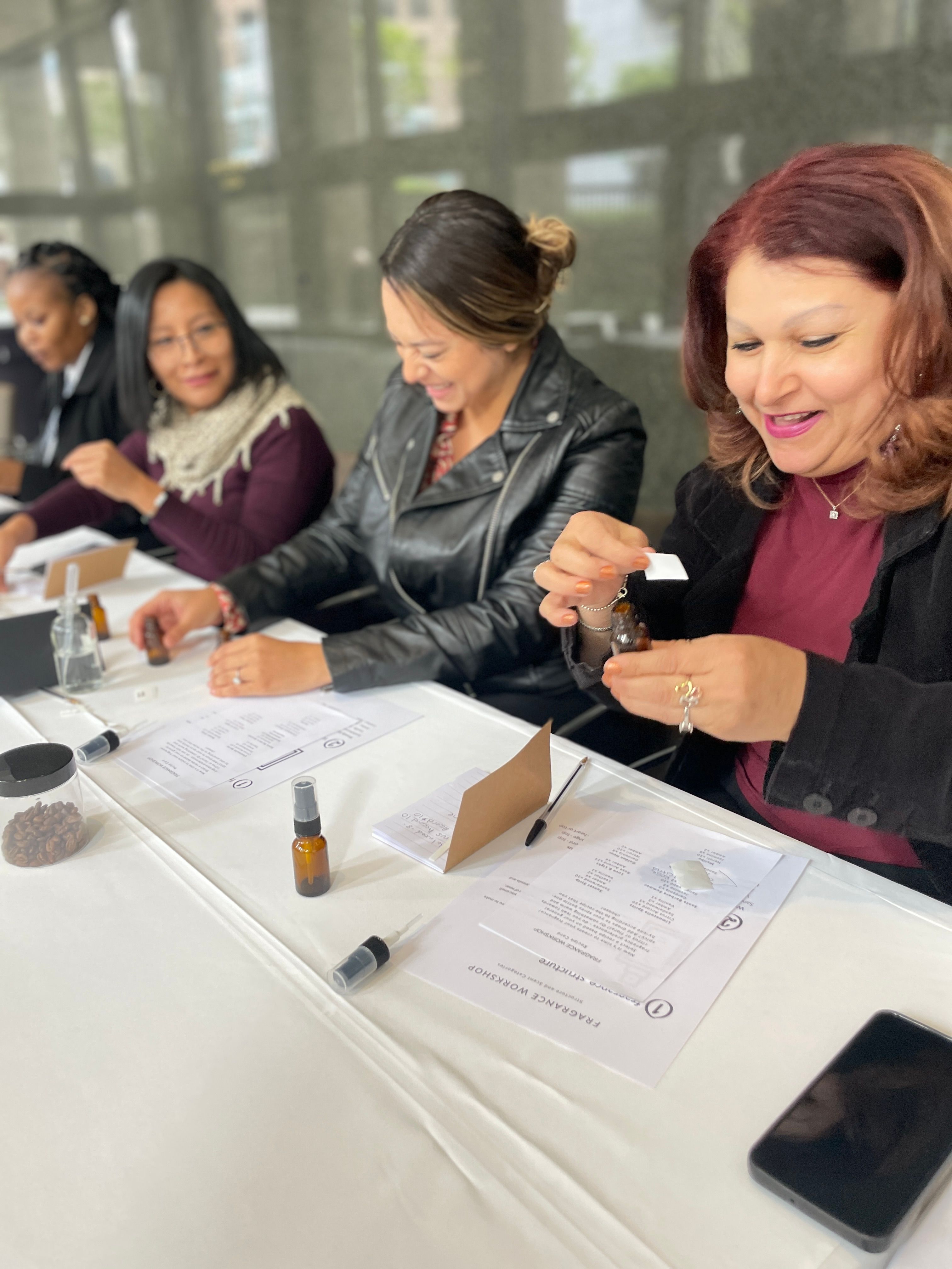 Smiling women at an indoor fragrance workshop mixing scents from small amber bottles over worksheets and notepads on a white table.