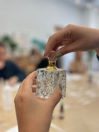 Close-up of hands with muted green nail polish removing a faceted stopper from an ornate crystal perfume bottle with a gold neck on a blurred workshop table