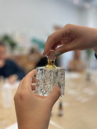 Close-up of hands with muted green nail polish removing a faceted stopper from an ornate crystal perfume bottle with a gold neck on a blurred workshop table
