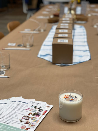 Cozy craft workshop table covered in brown paper with a blue-striped runner, a row of labeled kraft boxes down the center, glasses and pencils at each place, printed November activity sheets, and a decorative crystal-topped candle in the foreground.
