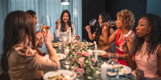 Group of women enjoying a cozy indoor dinner party, raising wine glasses in a candlelit toast around a table with a pink floral centerpiece.