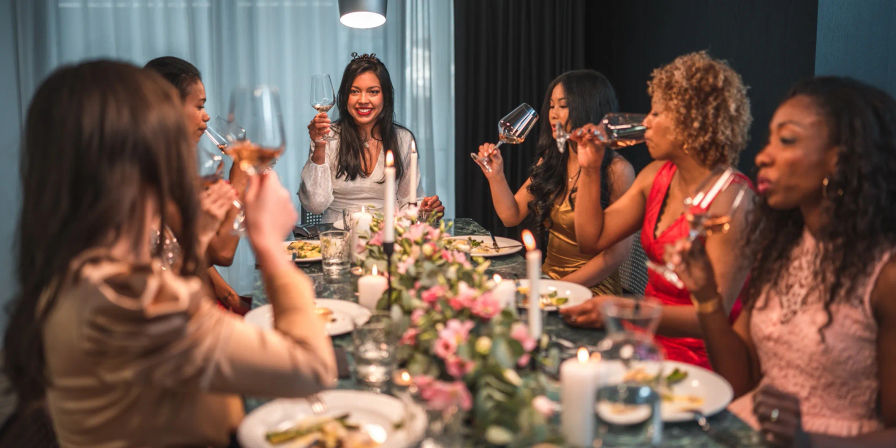 Group of women enjoying a cozy indoor dinner party, raising wine glasses in a candlelit toast around a table with a pink floral centerpiece.