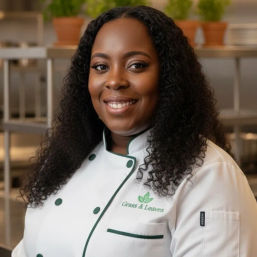 Smiling professional chef portrait — Black woman in a white double-breasted jacket with green trim, standing in a stainless-steel restaurant kitchen with potted herbs on shelves.