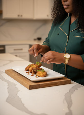 Chef's hands in a green jacket garnishing crispy fried croquettes with microgreens on a white rectangular plate set on a wooden board atop a white marble kitchen countertop