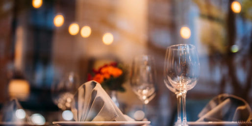 Cozy restaurant table with folded linen napkin on a plate, empty wine glasses, small floral centerpiece and warm bokeh lights — intimate city dinner setting.