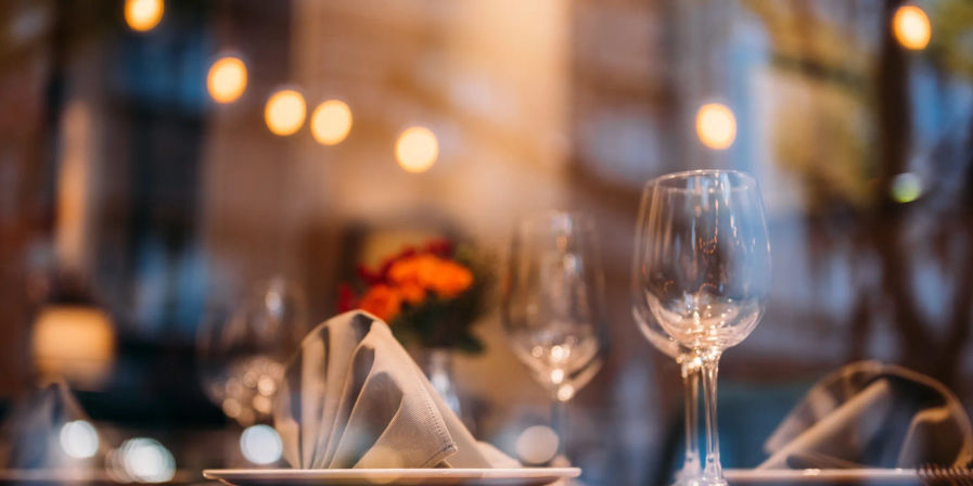Cozy restaurant table with folded linen napkin on a plate, empty wine glasses, small floral centerpiece and warm bokeh lights — intimate city dinner setting.