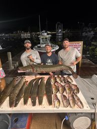 Three anglers on a dimly lit marina dock at night proudly holding a giant alligator gar above a cleaning table lined with multiple large gars and several smaller saltwater fish, with fishing boats and slips in the background.