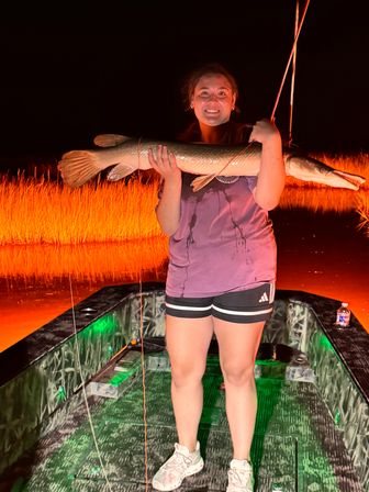Smiling angler holding a large pike-like fish on a small fishing boat at night with green deck lights and orange-lit marsh reeds in the background — big night fishing catch.