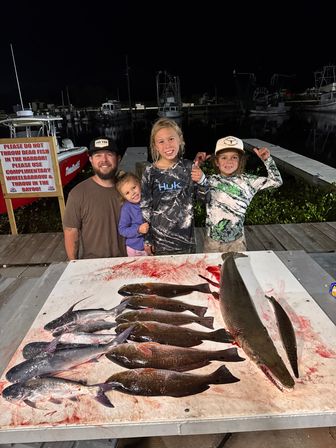 Nighttime marina scene with a family of four posing on a dock behind a table covered in a fresh catch of multiple fish, including a long-snouted gar; boats and pilings softly lit in the background.