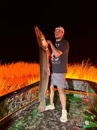 Night fishing: angler on a camo boat holding a giant alligator gar upright beside glowing marsh grasses