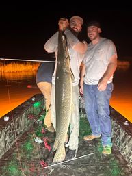 Night fishing on a small boat: two anglers proudly holding a giant alligator gar vertically over the deck, marsh grasses and orange-lit water glowing in the background.