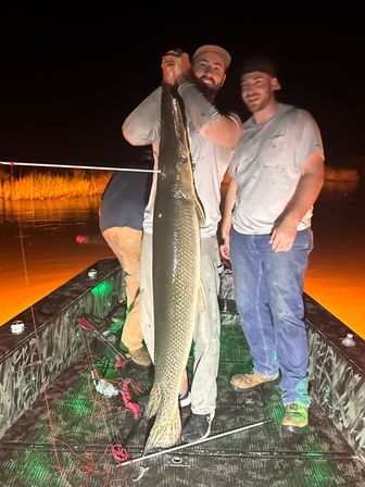 Night fishing on a small boat: two anglers proudly holding a giant alligator gar vertically over the deck, marsh grasses and orange-lit water glowing in the background.