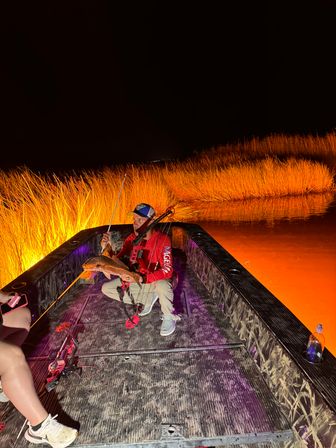 Night bowfishing scene: an angler kneels on a flat-deck boat holding a freshly caught fish as bright orange-lit marsh grasses and water glow behind, with bowfishing gear visible on the deck.