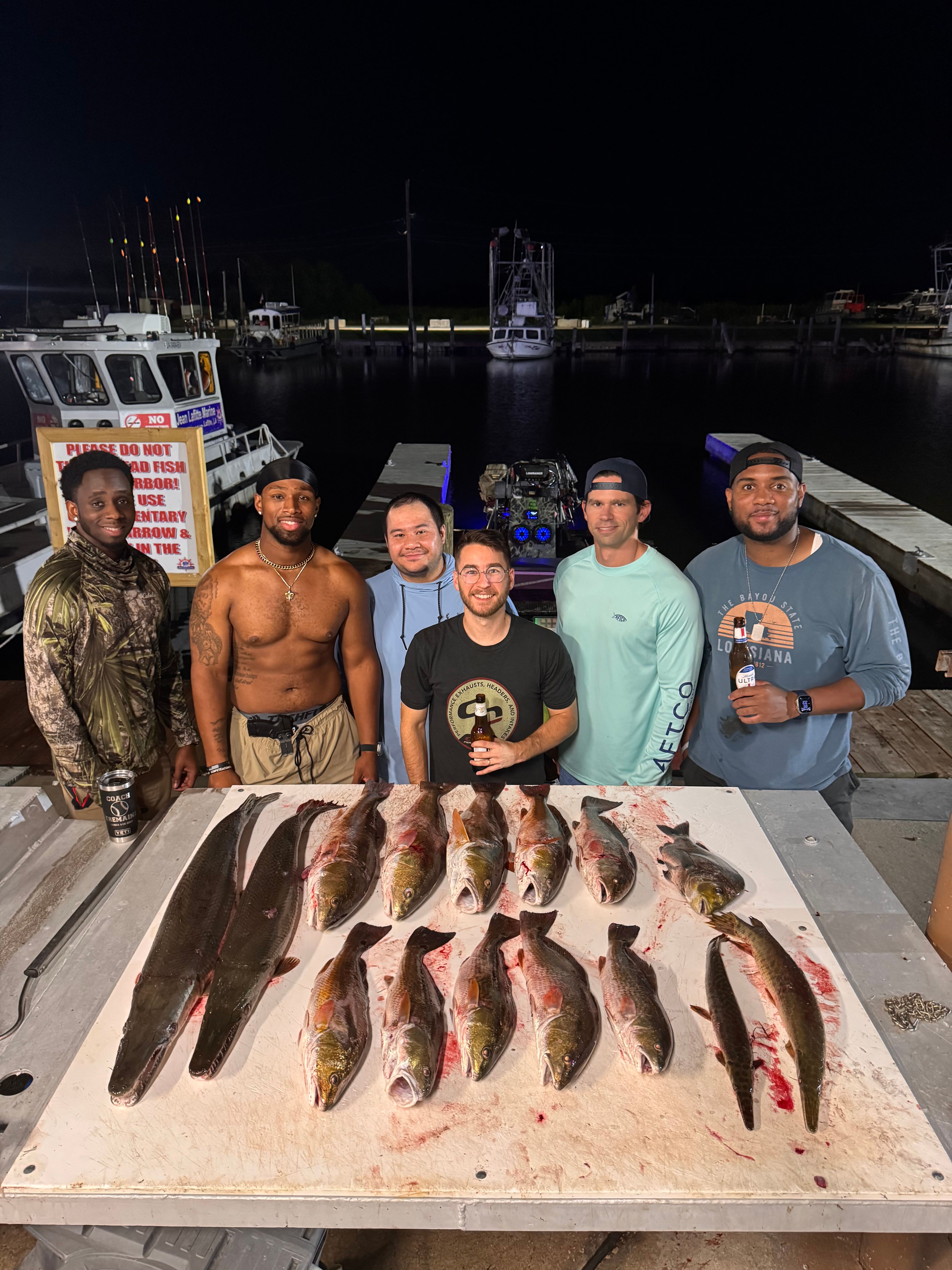 Six men pose on a dock at a coastal marina at night with a table of freshly caught saltwater fish laid out in rows, fishing boats and piers visible in the background.