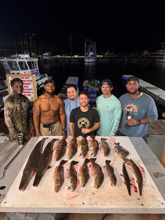 Six men pose on a dock at a coastal marina at night with a table of freshly caught saltwater fish laid out in rows, fishing boats and piers visible in the background.