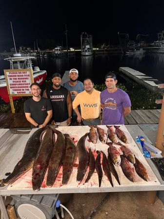 Five anglers posing behind a dockside cleaning table piled with a large nighttime catch — several long snouted fish and a row of medium-sized red-brown fish on a blood-streaked table, with boats and dark water at the marina in the background.