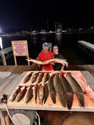 Night fishing dock scene: two anglers holding a large gar over a cleaning table with multiple gars and mixed saltwater catch, boats and harbor in the background.