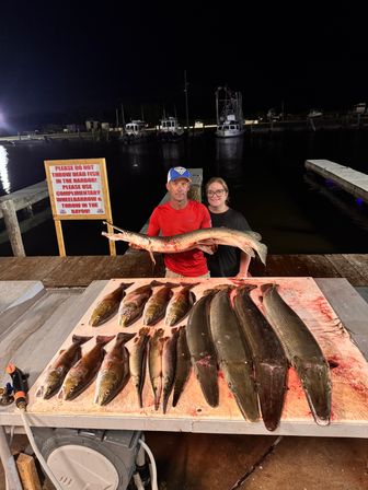 Night fishing dock scene: two anglers holding a large gar over a cleaning table with multiple gars and mixed saltwater catch, boats and harbor in the background.