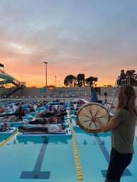 Sunset sound bath at an outdoor swimming pool with rows of people resting on floating mats in lanes while a facilitator plays a hand drum at poolside beneath a pastel sky.