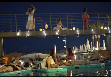 Nighttime pool sound-bath at a coastal resort — people relaxing on floating mats beneath a lit walkway where performers play a gong, candles and string lights glowing, palm trees and pool umbrellas in the background.