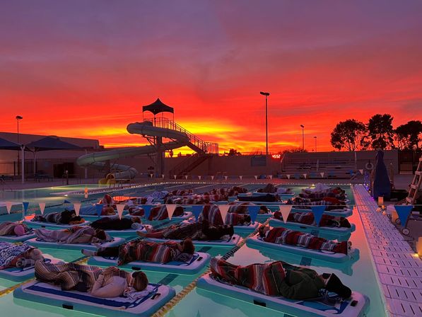 Cozy float-in at an outdoor pool: people wrapped in blankets on floating loungers beneath a fiery orange-red sunset, with a silhouetted waterslide and pool deck lights.
