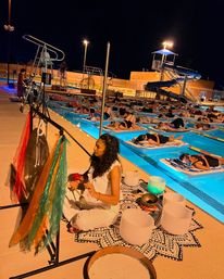 Nighttime outdoor pool sound bath at a community swimming facility — a facilitator on the deck plays gongs and crystal bowls while dozens relax on floating mats in glowing lanes near a waterslide.