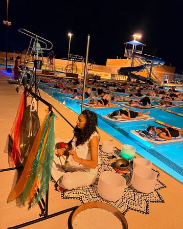 Nighttime outdoor pool sound bath at a community swimming facility — a facilitator on the deck plays gongs and crystal bowls while dozens relax on floating mats in glowing lanes near a waterslide.
