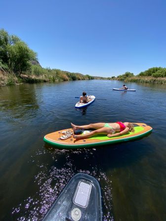 Sunlit summer river with three paddleboarders — one sunbathing on a colorful board, others paddling past reeds and green trees under a clear blue sky.