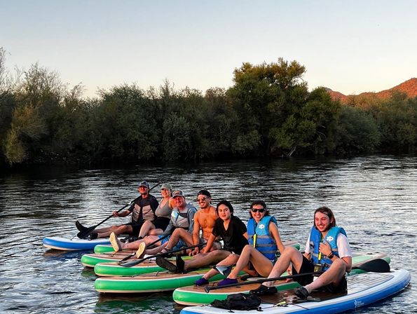 Group of seven people on inflatable stand-up paddleboards (SUPs) relaxing on a calm river at sunset, wearing life jackets and smiling with tree-lined banks and low hills in the background.