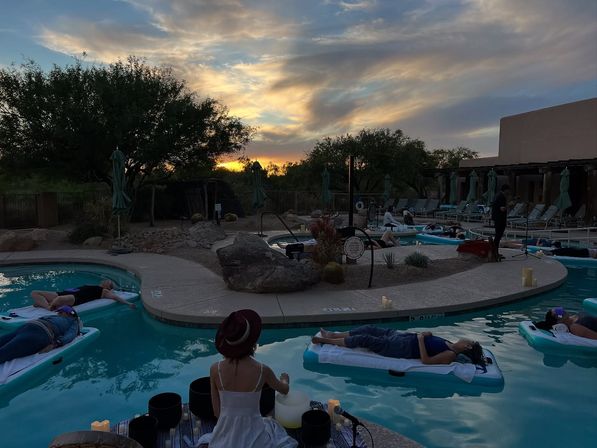 Desert resort pool at sunset with people relaxing on inflatable floats for a twilight sound bath, candles and lounge chairs around turquoise water and scrubby trees.