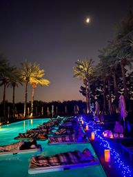Moonlit palm-lined pool at night with people resting on floating mats wrapped in striped blankets, turquoise underwater lights, blue fairy lights and glowing candles along the deck for a relaxed tropical resort vibe.