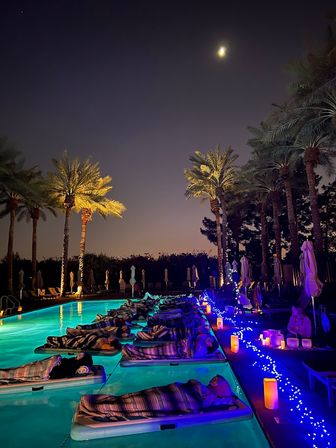Moonlit palm-lined pool at night with people resting on floating mats wrapped in striped blankets, turquoise underwater lights, blue fairy lights and glowing candles along the deck for a relaxed tropical resort vibe.