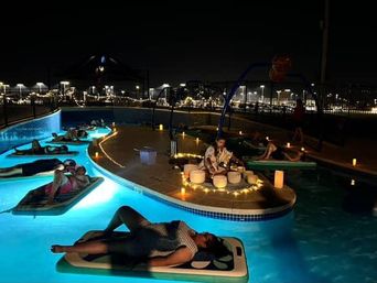 Nighttime outdoor pool sound-bath: people relax on floating mats in glowing blue water around a candlelit island where a musician plays crystal bowls, with string lights and city lights in the background.
