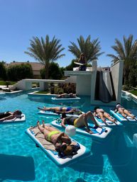 Group of people sunbathing on inflatable floating mats in a sunny resort-style backyard pool with palm trees and a waterslide