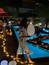 Nighttime pool sound bath — barefoot musician walks a string-lit pool edge with a frame drum as people relax on floating mats under blue pool glow and candlelight.