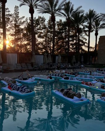 Sunset by a palm-lined outdoor pool with people on inflatable floats wrapped in blankets for a cozy floating movie night.