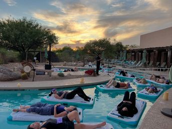 Guests relaxing on floating mats in a desert resort pool at sunset during a guided pool‑float meditation, surrounded by cacti, lounge chairs and soft evening light.