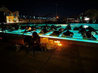 Nighttime outdoor pool sound bath with participants on glowing aqua-lane floats, candles and a musician playing a gong and crystal singing bowls for a relaxing group event.
