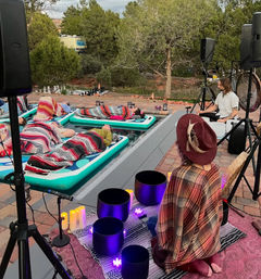 Outdoor poolside sound bath — people wrapped in colorful blankets lie on inflatable mats while facilitators play crystal singing bowls and a gong in a desert garden setting, candles and lanterns glowing for a cozy meditation session.