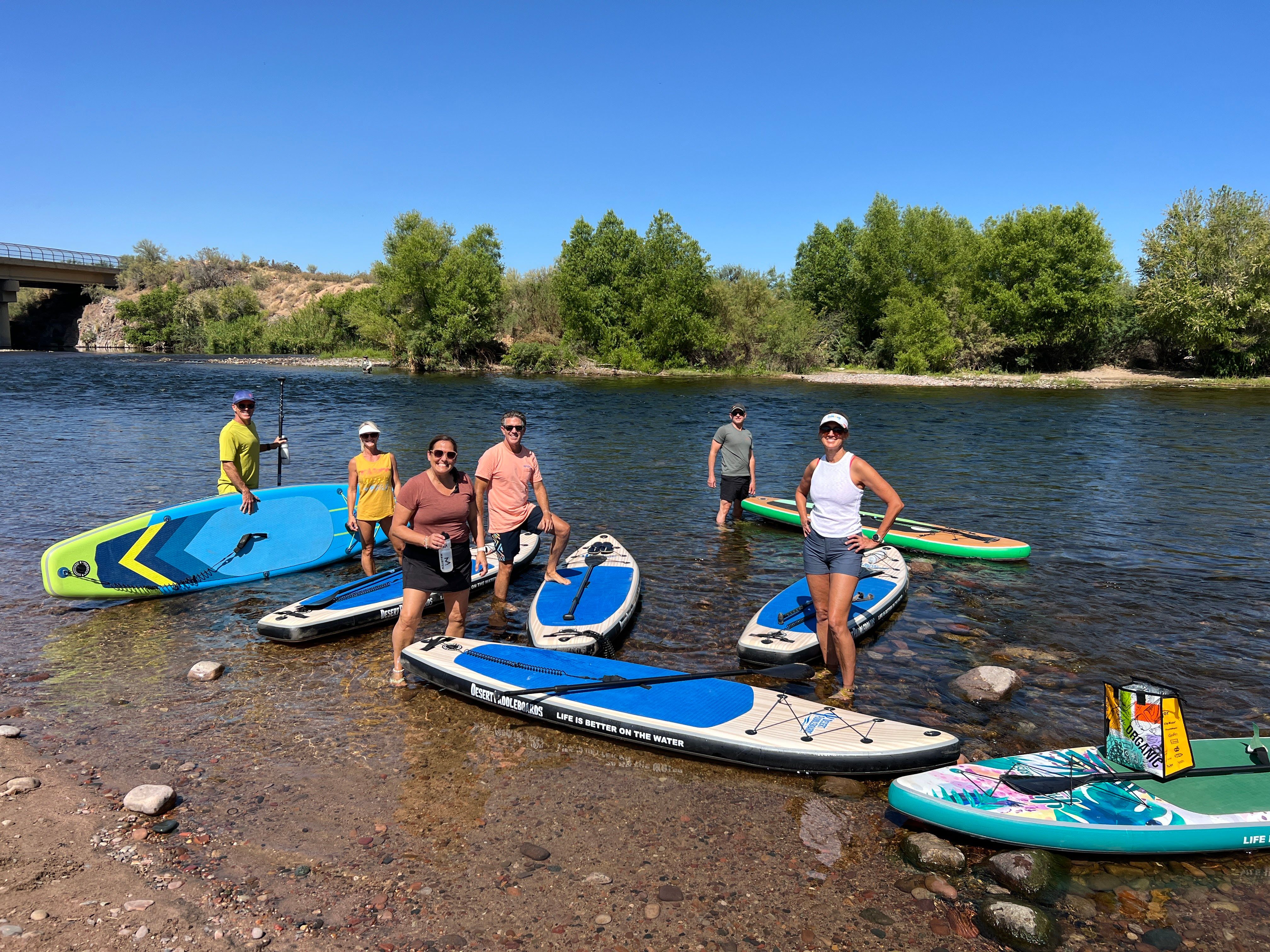 Six people with colorful stand-up paddleboards standing in shallow river near a tree-lined shore and bridge under a clear blue summer sky.