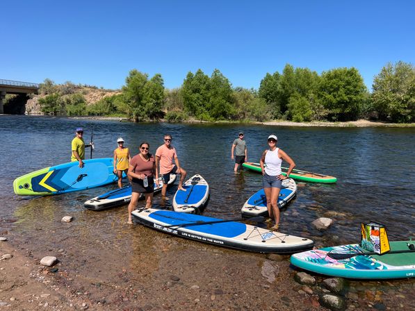 Six people with colorful stand-up paddleboards standing in shallow river near a tree-lined shore and bridge under a clear blue summer sky.