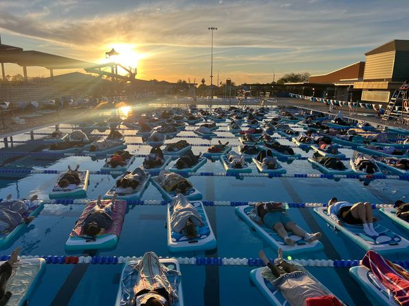 Sunrise community float at an outdoor lap pool — dozens resting on inflatable mats in lane lines with golden sky and a waterslide silhouette.