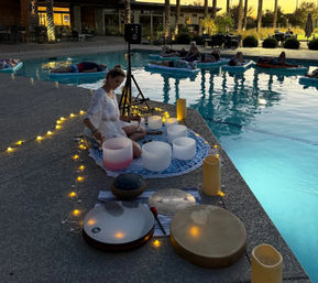 Evening poolside sound bath meditation: a person playing crystal singing bowls and hand drums on a mat beside a lit swimming pool while others relax on floating mats, string lights and candles creating a serene sunset atmosphere.