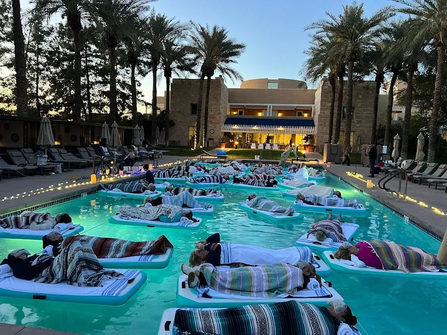 Evening pool float event at a palm‑lined resort — dozens of guests wrapped in colorful blankets on floating loungers in turquoise water, string lights and a warmly lit hotel building at dusk.