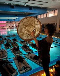 Candlelit sound bath at an indoor pool — a woman plays a large frame drum while participants rest on floating mats in glowing blue water during a relaxing floating yoga session.